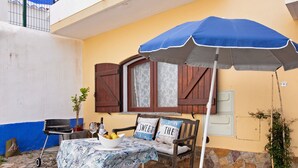 Une terrasse avec une table dressée pour le repas, un parasol bleu et un banc en bois agrémenté de coussins.