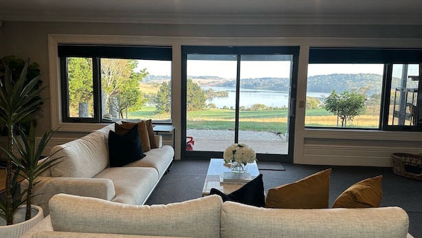 Un salon avec un canapé beige, une table basse et de grandes fenêtres donnant sur un lac et le paysage environnant.