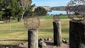 Un cadre extérieur paisible, avec des poteaux en bois soutenant des structures sphériques faites de branches entrelacées, le tout se détachant sur un paysage composé d’un lac, d’arbres et d’un ciel dégagé.