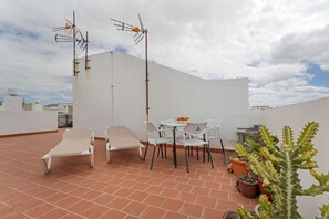 Una terraza en la azotea con sillas blancas, una mesa, plantas en macetas y dos antenas.