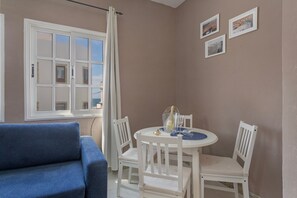A room with a blue sofa, a white dining table with chairs, a window with curtains, and framed pictures on the wall.