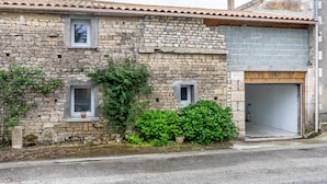 Un bâtiment en pierre avec deux petites fenêtres, un garage et un arbuste vert devant.