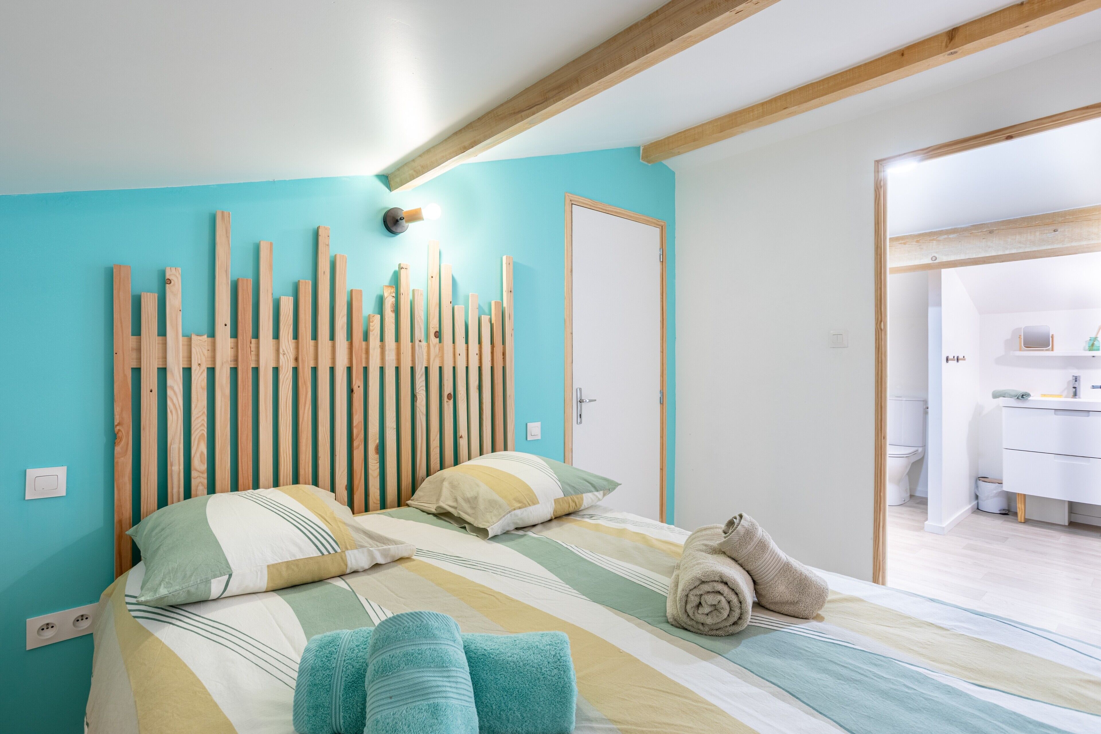 A bedroom with a wooden headboard, a bed with striped bedding, a rolled towel, and a view into the bathroom.
