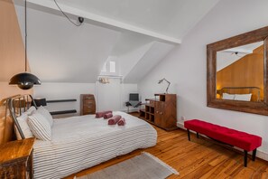 Um quarto com uma cama, um banquinho de madeira, uma escrivaninha, uma cadeira, um guarda-roupa e um espelho na parede.