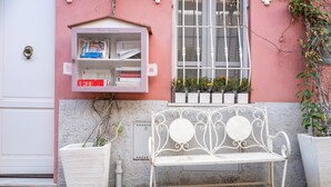 A bench outside a building with a pink wall, a window with white bars, and a small shelf with books.
