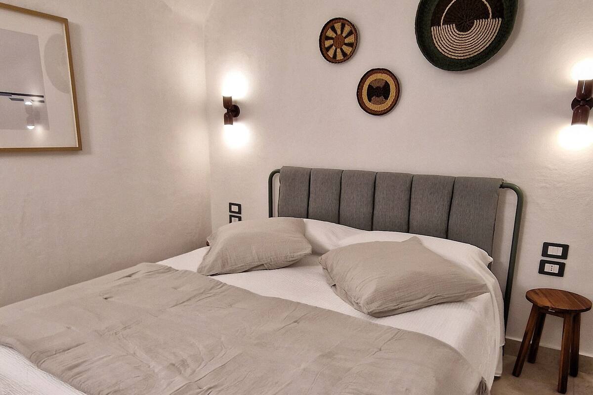 Ein ordentlich bezogenes Bett mit grauem Kopfteil, ein kleiner hölzerner Hocker und an der Wand befestigte dekorative Teller.