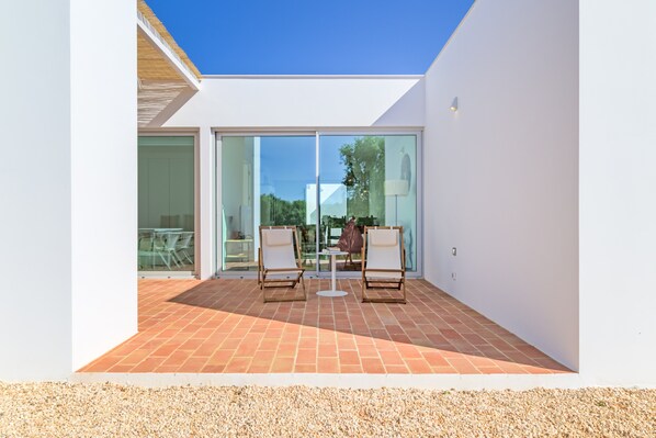 A modern outdoor patio with two chairs and a table, a glass door leading to an interior space, and a clear blue sky.