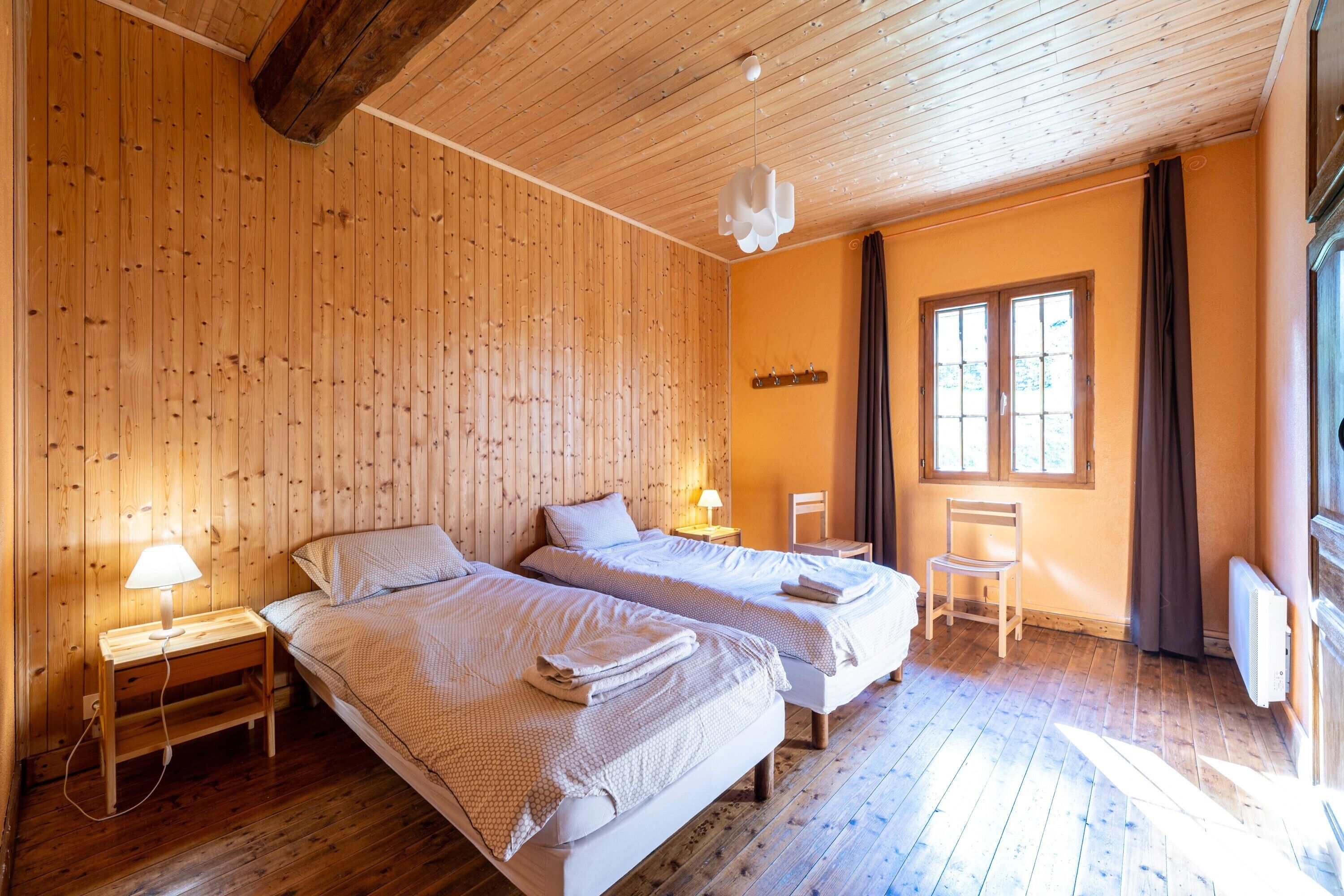 Ein Zimmer mit Holzwänden und -böden, zwei Betten, einem Sessel, einem Fenster und einer Lampe.