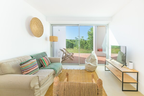 A modern living room with a sofa, a TV, a wooden bench, and a view through a glass door. - Holiday Home 'Casa Do Regadio - Casa 3' with Private Pool, Wi-Fi and Air Conditioning (Zambujeira do Mar)