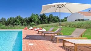 Zwembadgedeelte met ligbedden, een grote parasol en uitzicht op een huis en bomen.