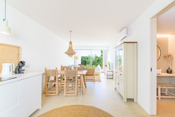 Une cuisine moderne avec des meubles blancs, un coin repas avec des chaises en bois, un luminaire suspendu et une vue dégagée grâce à de grandes fenêtres.