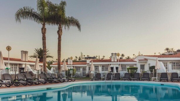 A poolside area with lounge chairs and umbrellas, palm trees, and buildings with red-tiled roofs. - 'Vista' with Shared Pool, Wi-Fi and Air Conditioning (Maspalomas)
