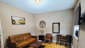 Una sala de estar con un sofá marrón, una mesa de centro, un escritorio con una silla, una lámpara y un televisor en la pared.