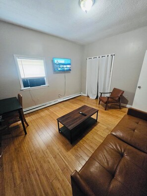 A living room with a brown leather couch, a coffee table, a TV on the wall, a window with blinds, and a door. - Nice spacious room near Whole Foods, Brown Univer. (Providence)