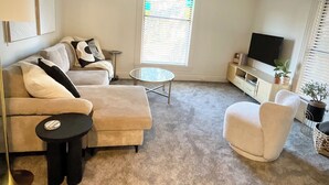 A living room with a beige sofa, a round coffee table, a small black table, a white armchair, a TV on a white cabinet, and a window with blinds. - Next Level Convenience in Grand Rapids premier neighborhood (Grand Rapids)