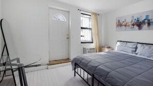 Un dormitorio con cama, una mesita de cristal, una puerta blanca, una ventana con cortinas y un cuadro de un paisaje urbano.
