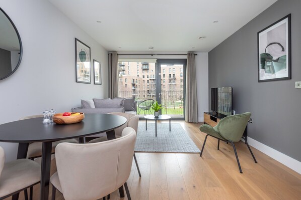 Una sala de estar moderna con una mesa de comedor, sillas, un sofá, un televisor y un balcón con vista a edificios.