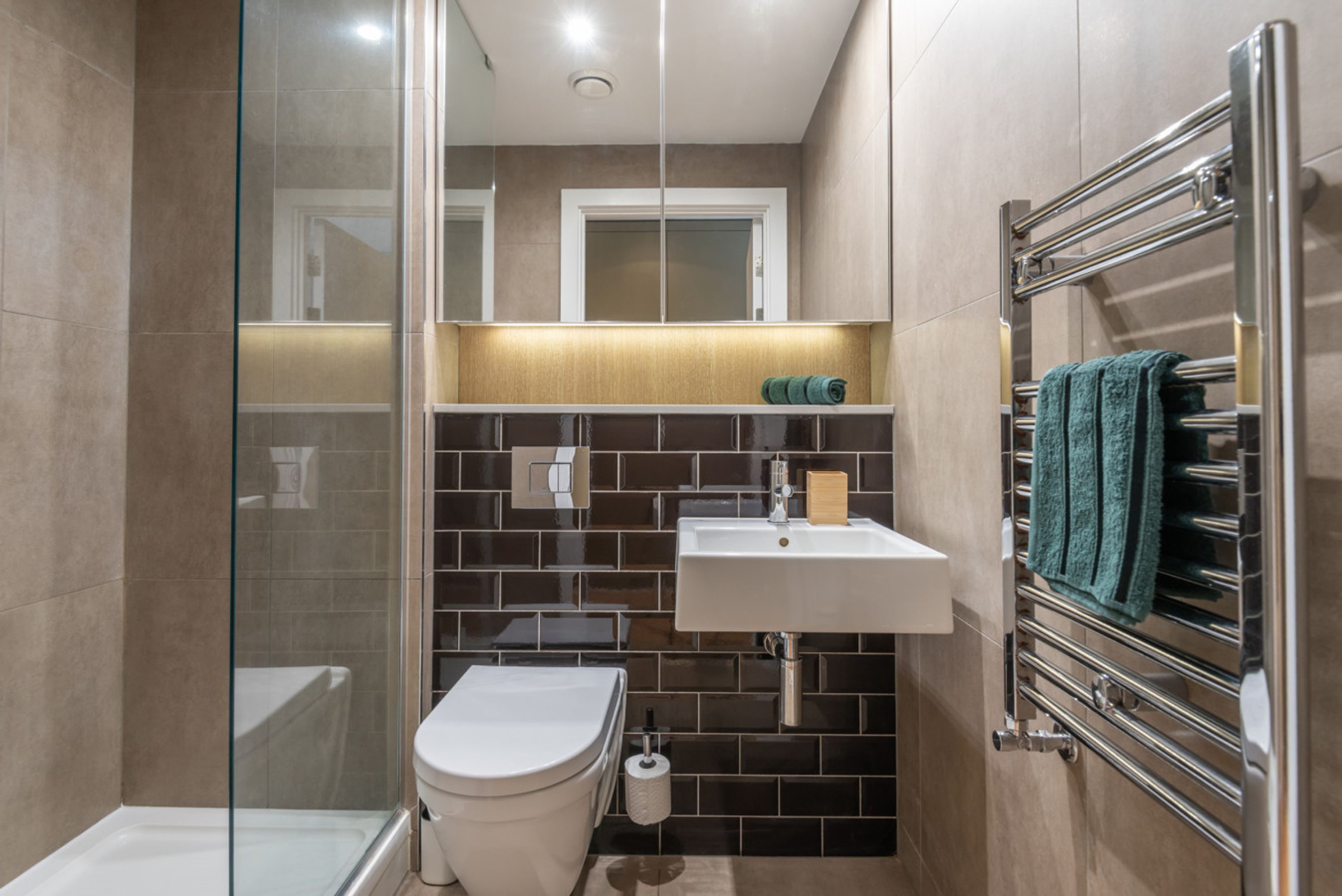 A bathroom with a glass shower enclosure, a white toilet, a sink with a mirror above, and a towel rack with a green towel.