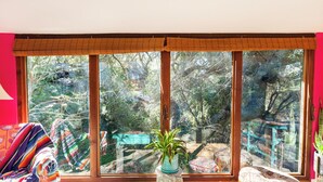 A room with large windows, a small stool, a chair, and a colorful blanket.