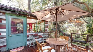 A wooden deck with a table and chairs, a large umbrella, and a rustic cabin-style building with a green door.