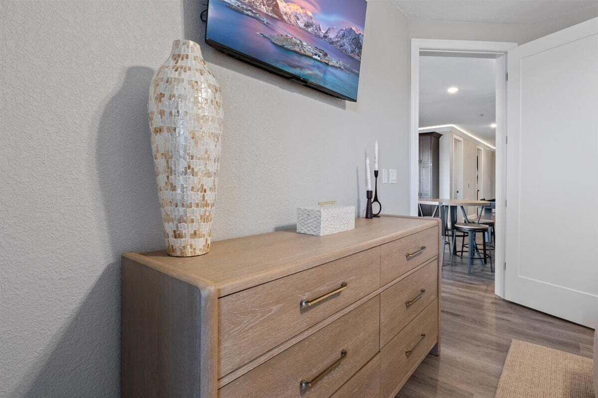 Un armari de fusta amb calaixos, un vas mosaic i un televisor que mostra un paisatge idíl·lic, i una porta que dóna a una zona de menjador.