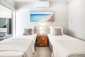 A hotel room with two beds, a nightstand with a lamp, and a beach-themed painting on the wall.