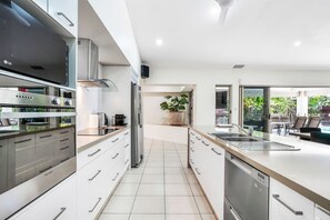 Una cocina moderna con horno empotrado, refrigerador y lavavajillas. Se ve un comedor con asientos exteriores a través de las puertas de vidrio.