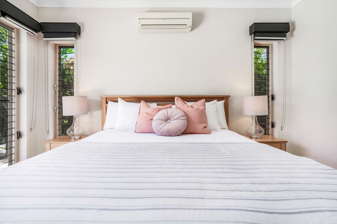 A neatly made bed with striped bedding, two bedside lamps, and a window with blinds on either side.