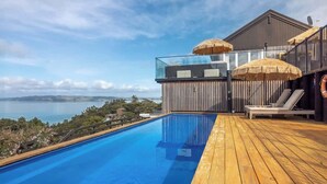 A modern infinity pool with a wooden deck, lounge chairs, and thatched umbrellas, overlooking a scenic landscape. - Woodside Bay Estate (Waiheke Island)
