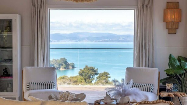 A room with two white chairs facing a large window overlooking a body of water and distant land. - Woodside Bay Estate (Waiheke Island)