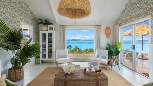 A spacious living room with a large window offering a view of the sea, a wicker coffee table, white sofas, and tropical plants. - Woodside Bay Estate (Waiheke Island)