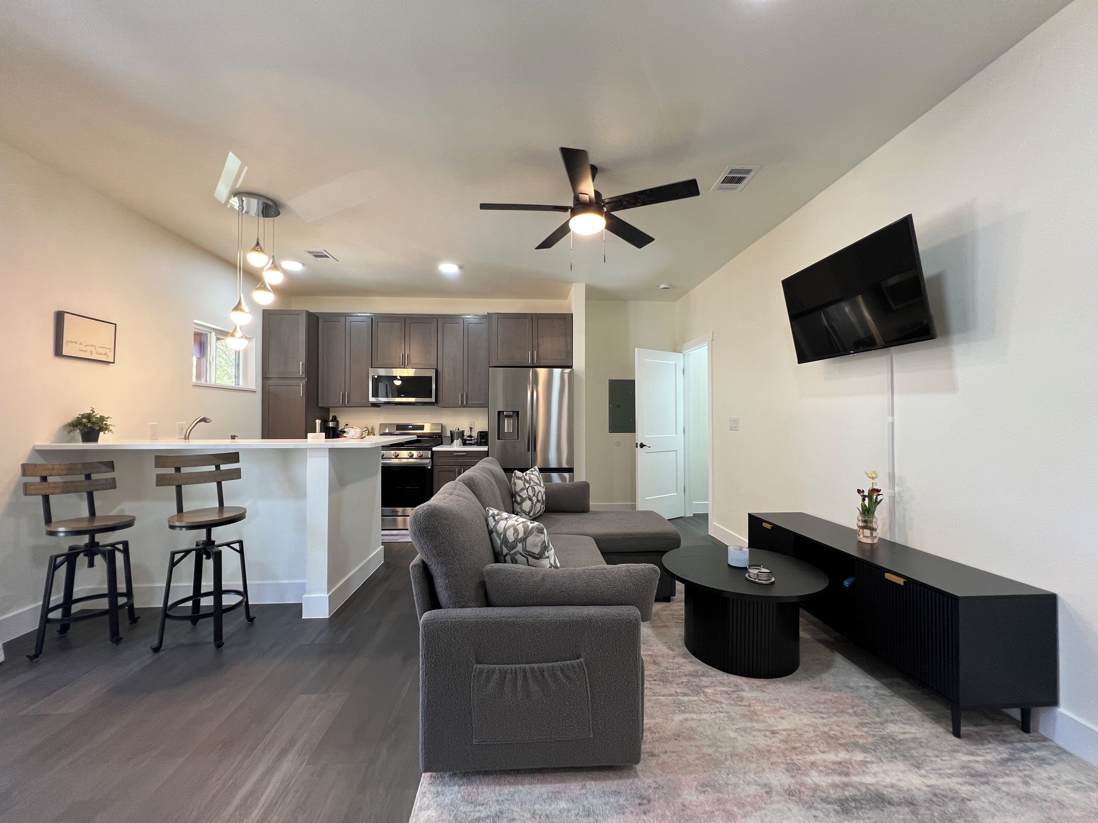 A modern kitchen with dark cabinets, a central island with stools, a dining area with a gray sofa and coffee table, and a flat-screen TV mounted on the wall.