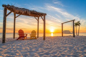 Escena de playa con una estructura de madera, dos sillas Adirondack y un columpio.