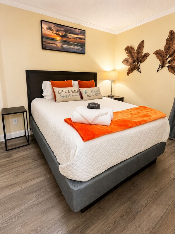 Una cama bien hecha con una colcha blanca y naranja, un almohadón con texto y un objeto negro pequeño sobre la cama.