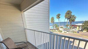 En balkon med to fletstole og et lille rundt bord, med udsigt over en strand med palmer og et poolområde.