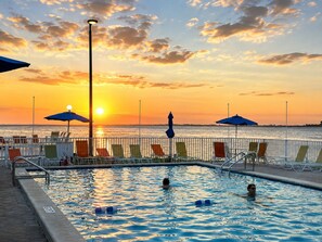 Una piscina con sillas de descanso y sombrillas, con vista a una puesta de sol sobre el agua.