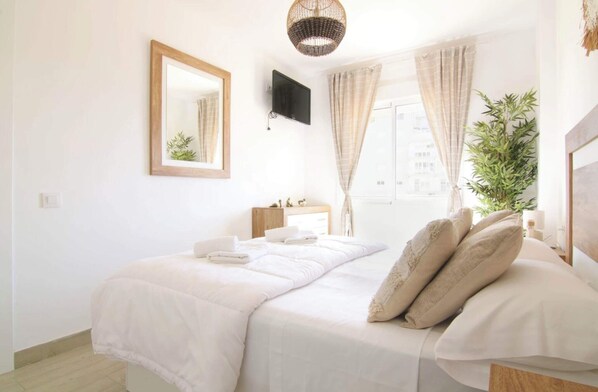 Un dormitorio ordenado con cama, mesita de noche, espejo, televisor y ventana con cortinas.