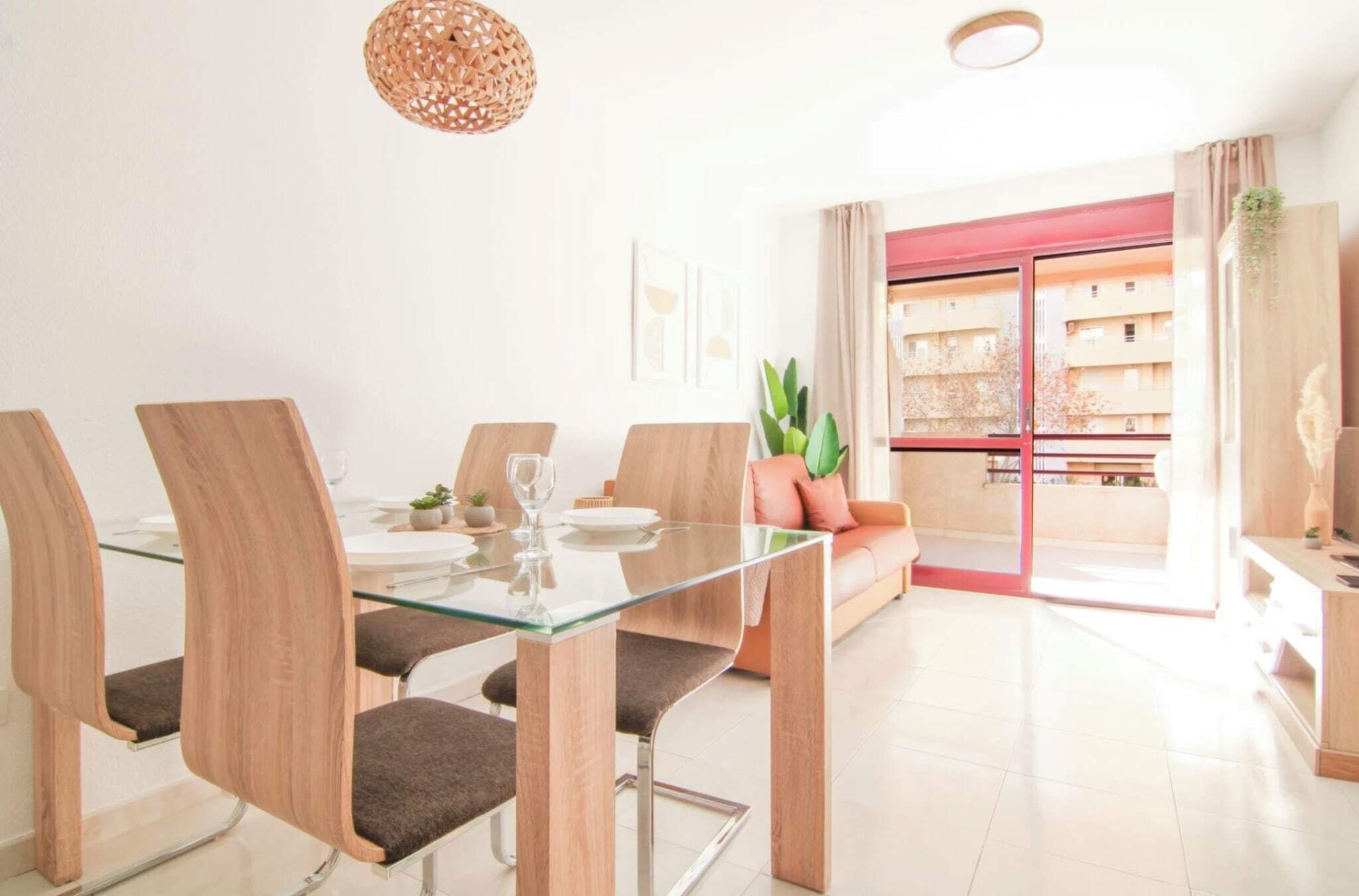 Un comedor moderno con una mesa de vidrio y sillas de madera, una planta en maceta y vistas a un edificio a través de un ventanal.