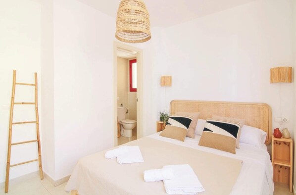 A neatly arranged bedroom with a bed, a bedside table, a lamp, and a bathroom visible through an open door.