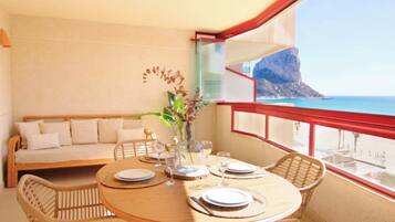 Un balcón con mesa de madera dispuesta para comer, un sofá de mimbre y vistas al mar y un acantilado.