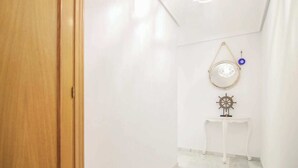 En hall med en trädörr, en vit vägg, ett litet vitt bord med en skeppsrat som dekoration och en rund spegel med en blå detalj.
