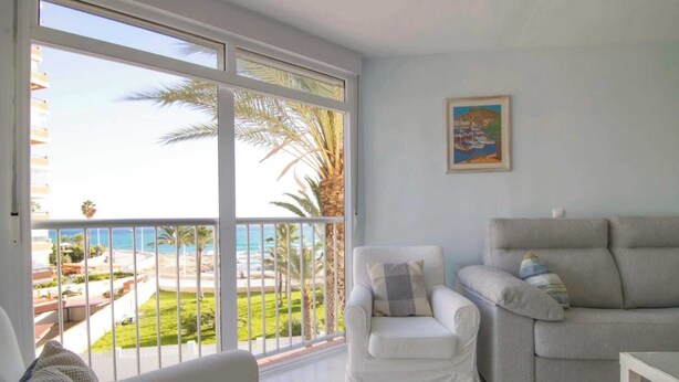 A living room with a view of the ocean, a white sofa, a white armchair, and a painting on the wall.