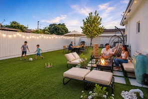A backyard with artificial grass, outdoor furniture, a fire pit, and string lights.