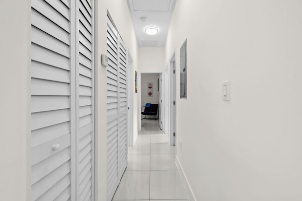 Un pasillo estrecho, con paredes blancas y suelo de baldosas, que conduce a una habitación con cama y escritorio.