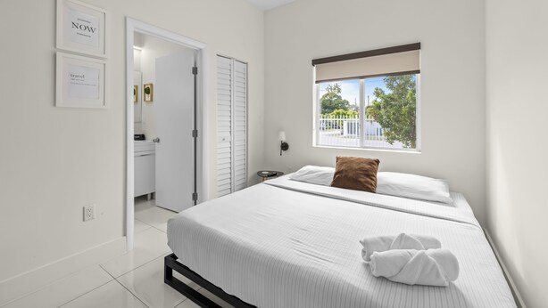 A modern bedroom with a large bed, a window with a view, and a door leading to a bathroom. - Family & Friends Fun-Huge Yard -Games -BBQ-FirePit (Miami)