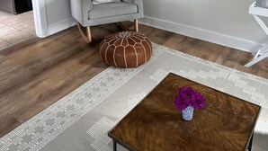 Una sala de estar con un sillón gris, un reposapiés marrón, una mesa de centro de madera con un jarrón de flores y una alfombra estampada.