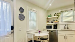 Cocina pequeña con gabinetes blancos, microondas, lavamanos y una zona de comedor con dos sillas.