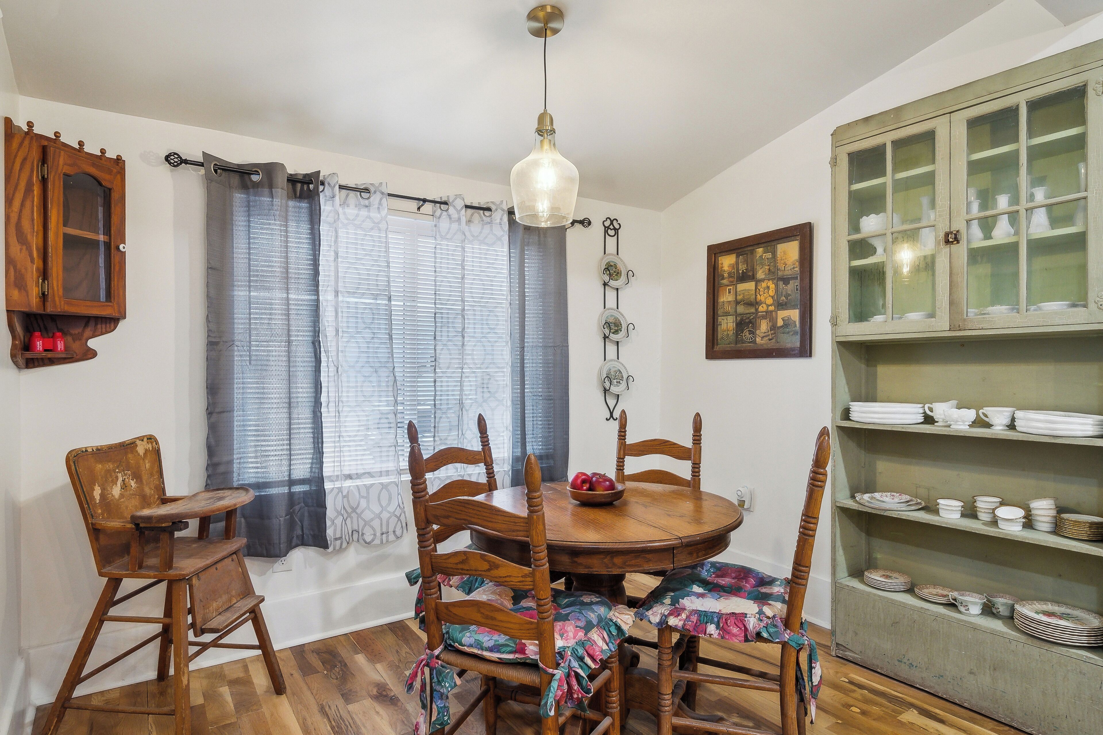 A dining area with a round wooden table, six chairs, a hanging light fixture, a window with curtains, and a cabinet with dishes.
