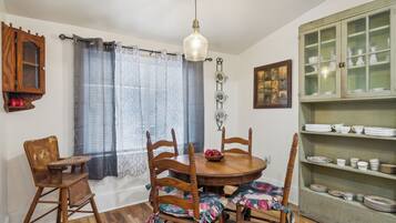 A dining area with a round wooden table, six chairs, a hanging light fixture, a window with curtains, and a cabinet with dishes.