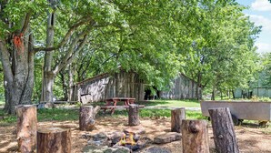 A rustic outdoor setting with a wooden fire pit surrounded by large rocks and tree stumps, a wooden bench, and a picnic table in the background. - 1800s Farmhouse—King Bed~Big Kitchen~Games & DVDs (Broken Arrow)